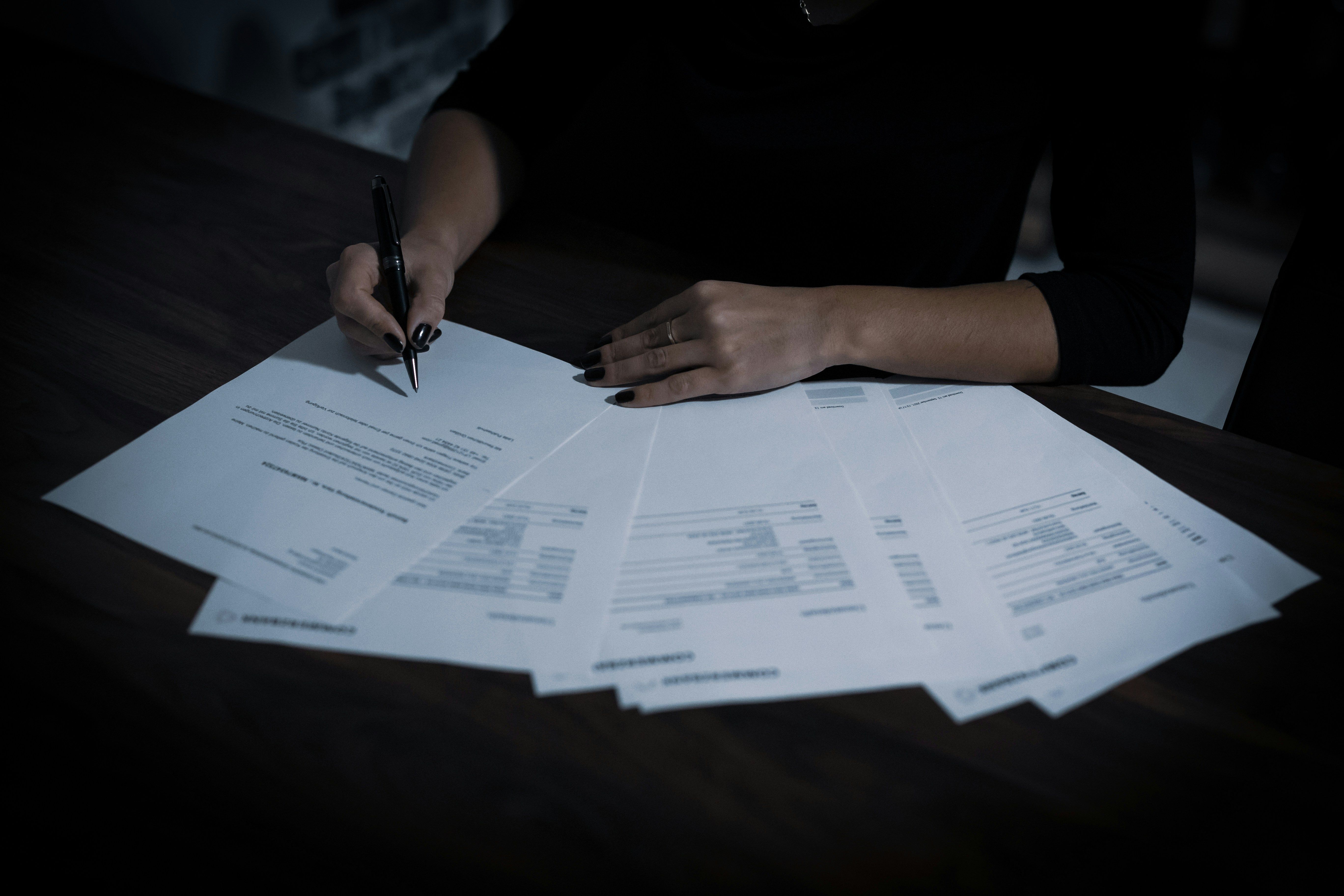 Close-up of a person signing important legal documents, with several papers neatly spread across a wooden desk. The image reflects the formal process involved in property transactions, from compliance checks to contracts. Dark tones create a professional and focused atmosphere, highlighting the importance of accuracy and attention to detail in estate agency and conveyancing work