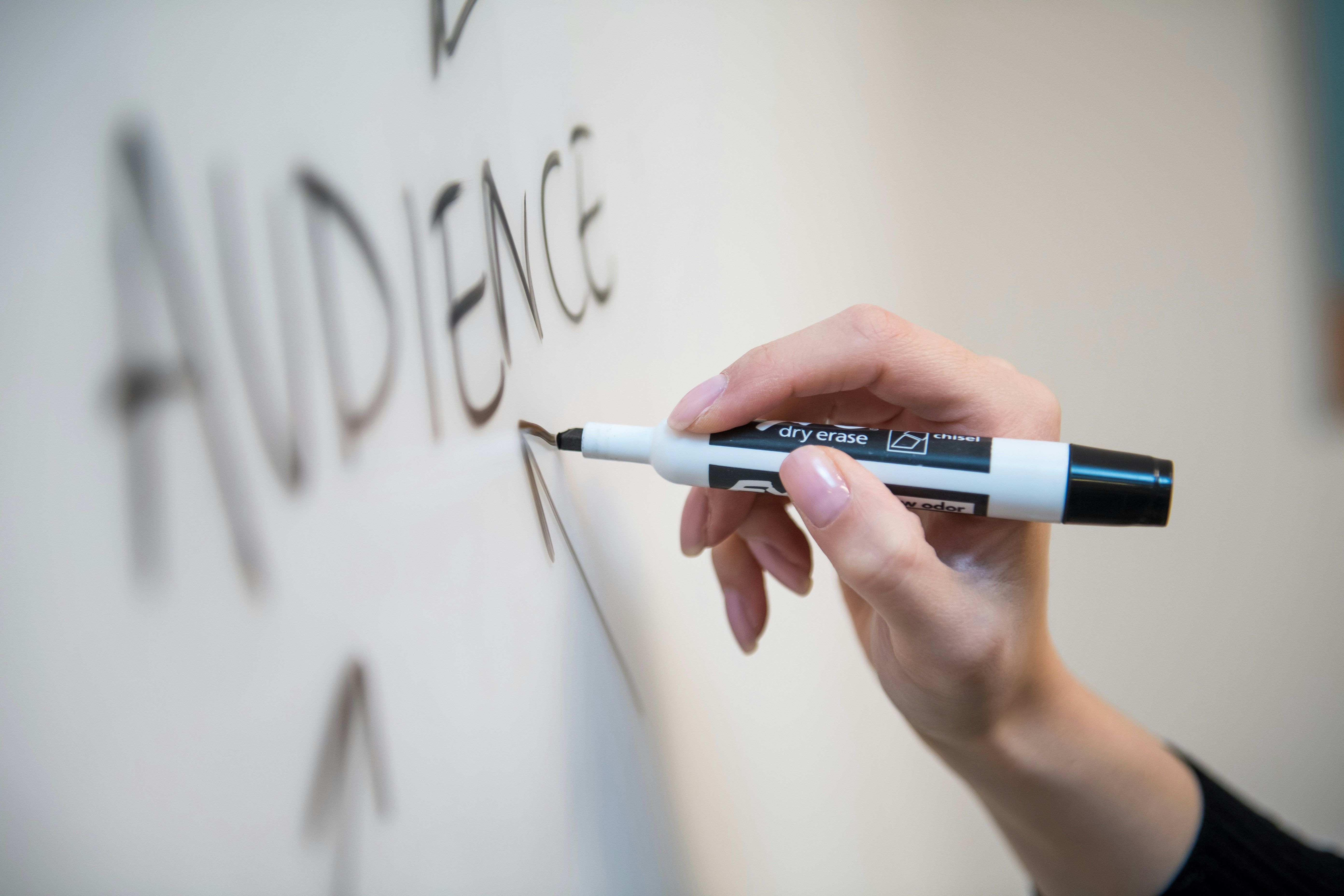 Close-up of a hand writing the word “Audience” on a whiteboard with a black dry-erase marker. The image highlights planning, communication, and strategy — key elements when identifying and targeting the right audience. Perfect for illustrating concepts such as marketing, presentations, or business planning, the photo captures focus and precision in action.