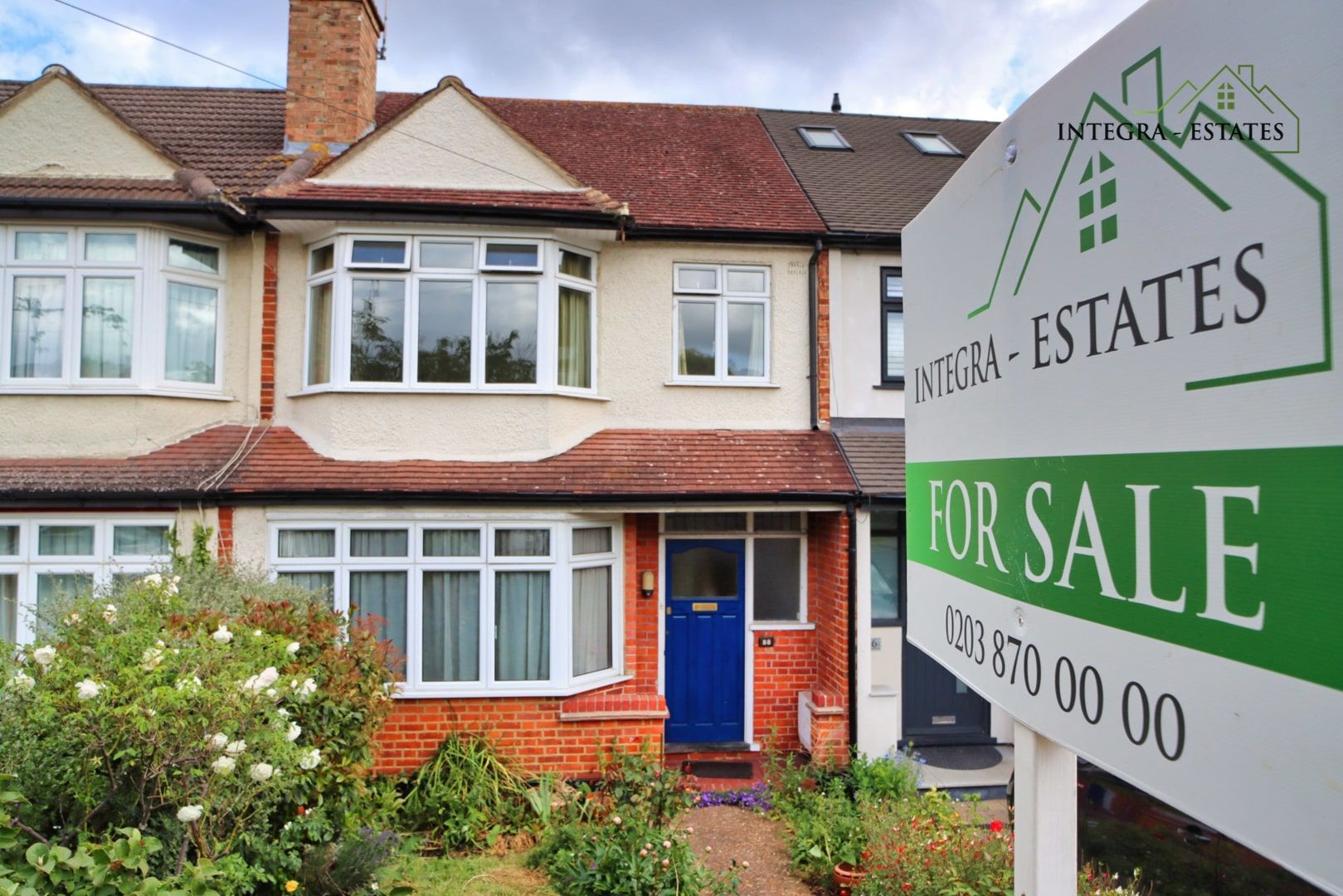 A semi-detached house with a red brick lower level and cream upper facade featuring large bay windows and a blue front door. A green and white Integra Estates “For Sale” sign is displayed prominently in the front garden, which is filled with plants and flowers. The photo represents a property listing in a suburban South East London or North Kent area.