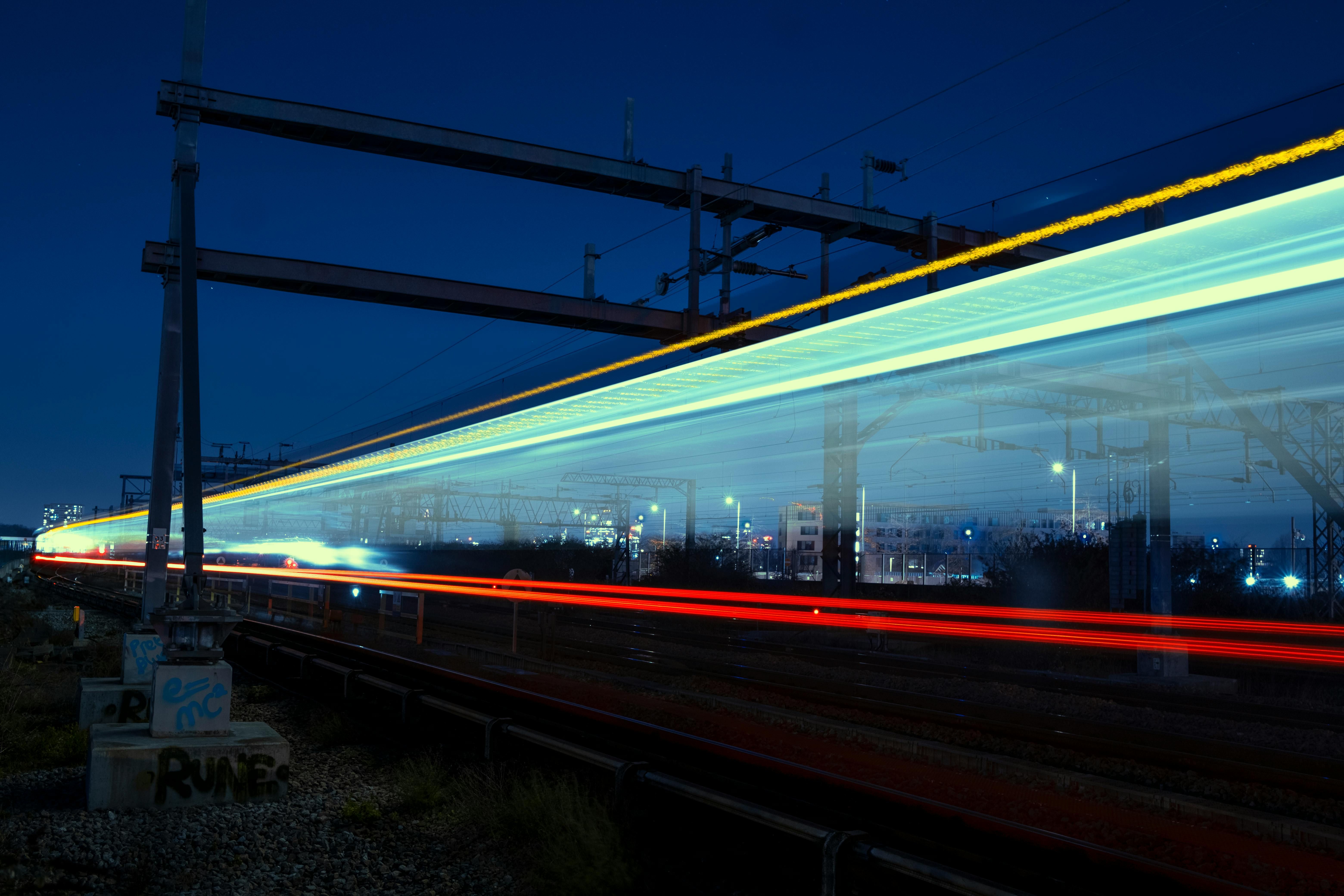 Night-time long-exposure image showing the motion blur of a train travelling along electrified railway tracks. The bright streaks of light in red, yellow, and blue highlight the speed and efficiency of urban transport. Overhead power lines, supporting structures, and distant city lights frame the scene, making it a striking representation of modern London transport and commuter connections across South East London.
