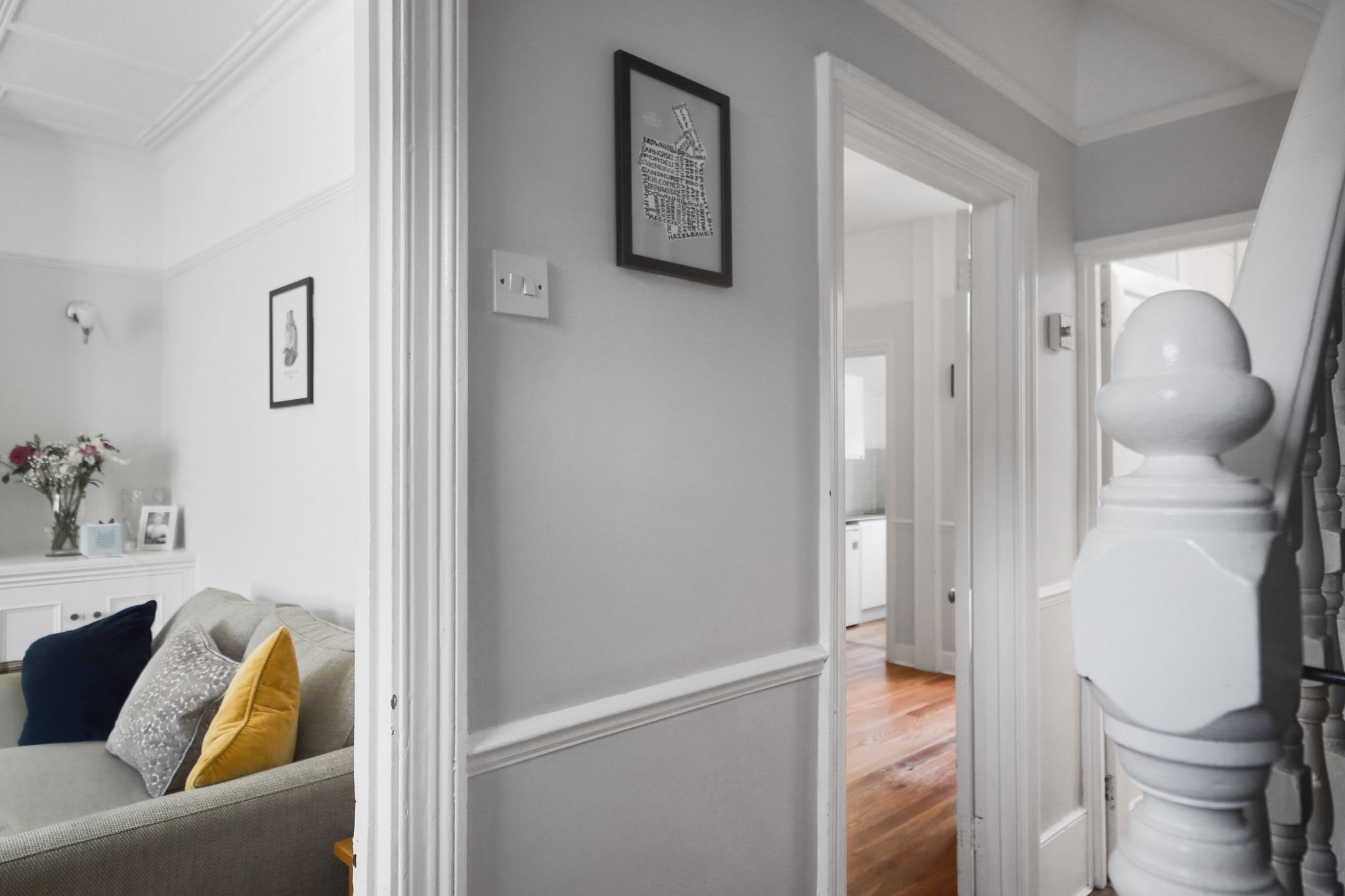 A bright and inviting hallway leading into a modern UK home, finished in soft neutral tones with clean white walls and natural wood flooring. The space opens onto a cosy lounge styled with a sofa, bold accent cushions, and fresh flowers, creating a homely feel. Classic staircase detailing and framed artwork on the walls add a touch of character, while natural light enhances the welcoming atmosphere, making this property ideal for comfortable family living.