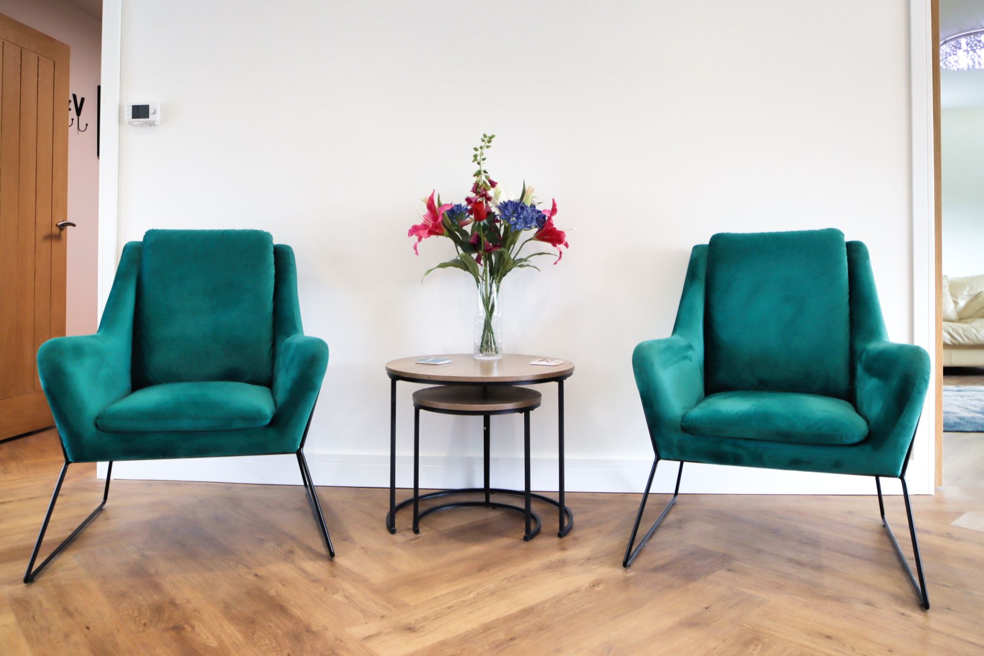 A stylish seating area with two modern emerald-green armchairs set against a crisp white wall. A sleek nesting side table sits between them, topped with a vase of fresh flowers that adds colour and elegance to the space. The warm wooden herringbone flooring enhances the contemporary feel, creating a welcoming corner perfect for relaxing or entertaining.