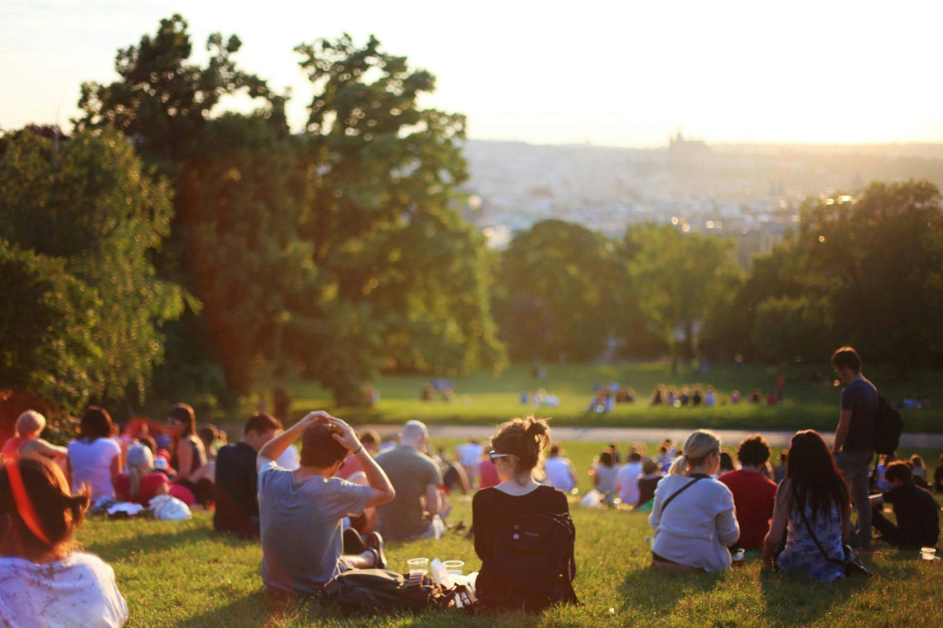 Vibrant summer scene in Orpington Park with people relaxing on the grass and soaking up the evening sun. Groups of families, friends, and couples sit together across the open green space, enjoying the warm weather and natural surroundings. Tall trees frame the view, while the distant city skyline fades into the golden glow of sunset. This image captures the sense of community, leisure, and relaxation that local parks in Orpington and South East London provide, making them attractive to families, young professionals, and property buyers looking for green spaces nearby. Perfect for SEO content about Orpington parks, outdoor living, and community lifestyle.