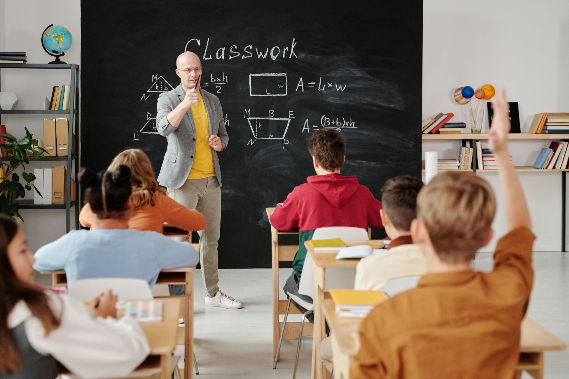Modern classroom in Orpington with a teacher delivering a maths lesson to engaged pupils. The teacher stands at the blackboard where geometric formulas and diagrams are written, while children sit attentively at their desks, with one pupil raising a hand to answer. Bright, organised shelves filled with books and a globe highlight the learning environment. This image reflects the quality of education, dedicated teachers, and positive learning atmosphere found in Orpington’s schools—an important factor for families moving to the area. Ideal for SEO targeting Orpington schools, family-friendly areas, and education in South East London.