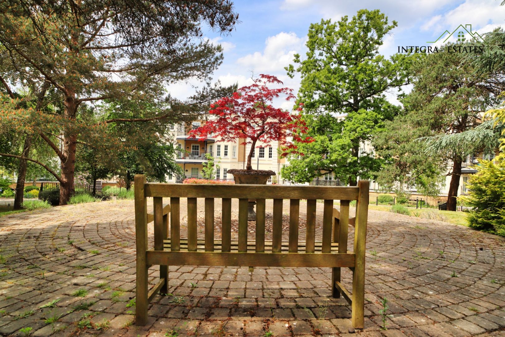 Beautiful landscaped outdoor space featuring a wooden bench overlooking a vibrant red maple tree in a circular courtyard, framed by tall green trees and modern residential buildings. This calm, well-maintained setting reflects the importance of outdoor living spaces in property developments, adding lifestyle appeal and enhancing property value. Perfect for SEO targeting outdoor space in Orpington homes, landscaped gardens in South East London, and properties with communal green areas.