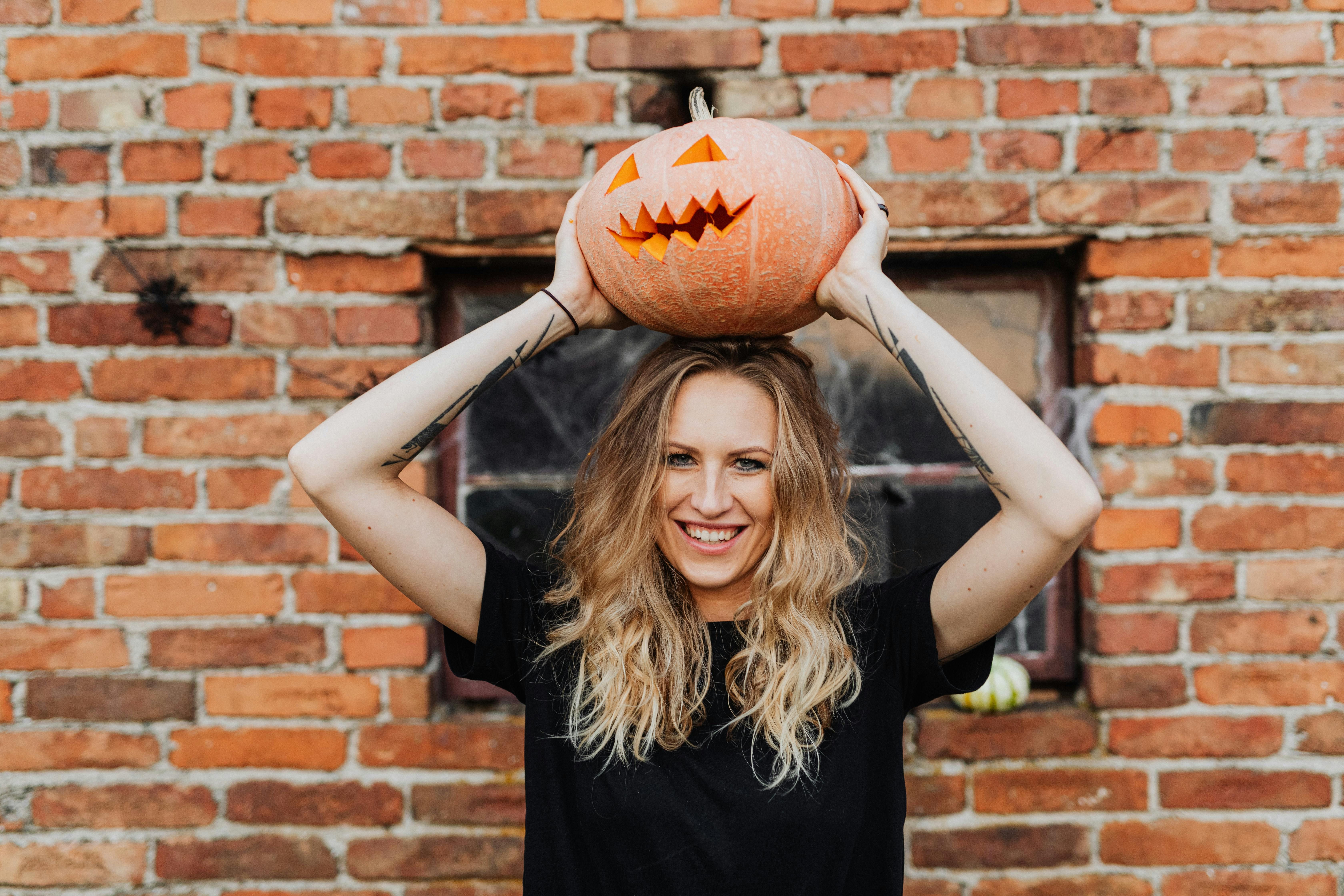 Young woman with long blonde hair smiling while lifting a carved jack-o’-lantern pumpkin above her head against a rustic brick wall backdrop. The image captures the fun and festive spirit of Halloween, making it ideal for seasonal blogs about community events, family activities, and neighbourhood celebrations in Beckenham and South East London