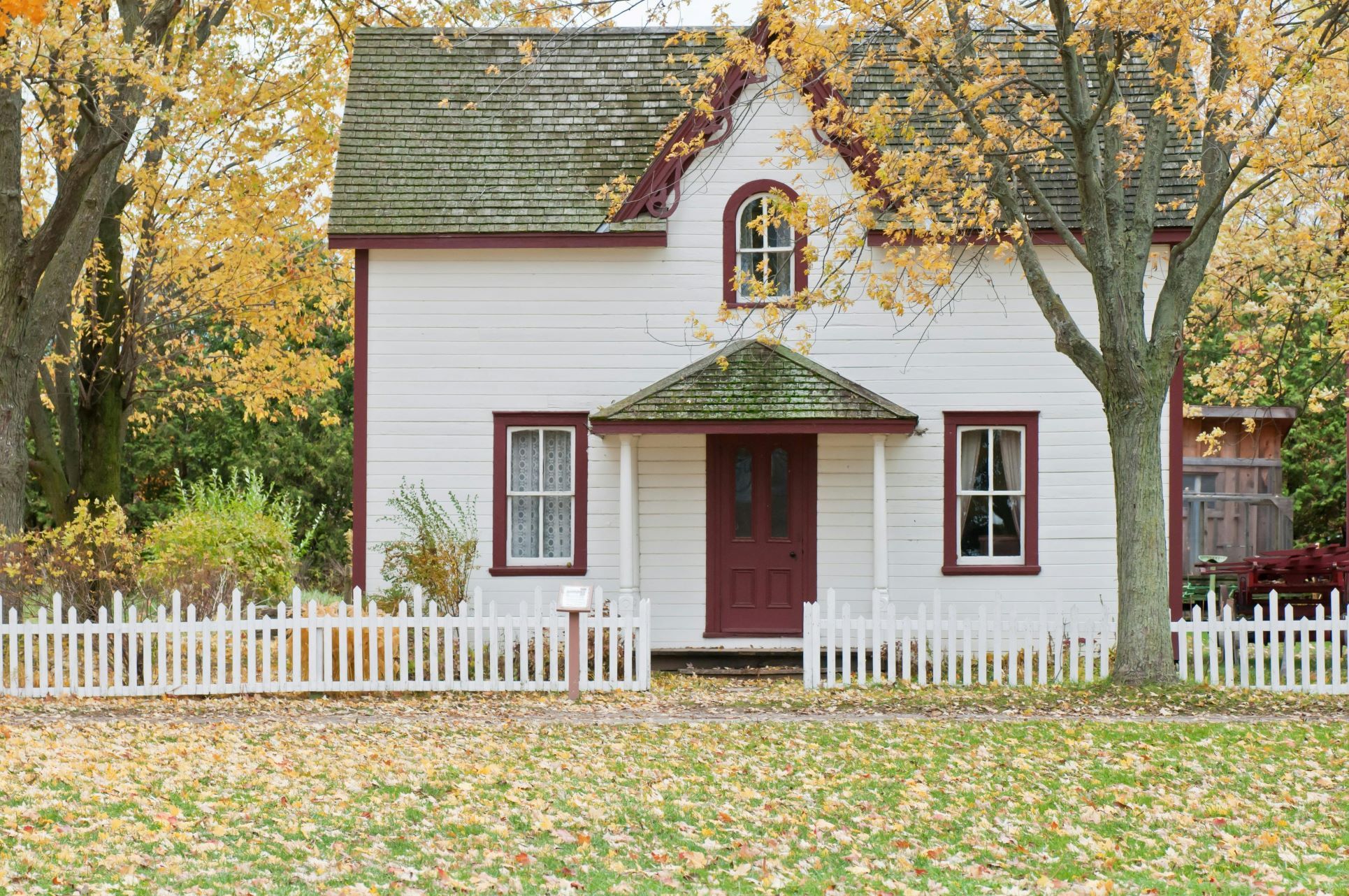 A quaint countryside-style home with white wooden cladding, red-framed windows, and a red front door, complete with a white picket fence. The property is framed by golden autumn leaves and trees, evoking a peaceful, homely, and nostalgic atmosphere. Ideal imagery for blogs about probate properties, remote living, property valuations, or homes with character in South East London and Kent.