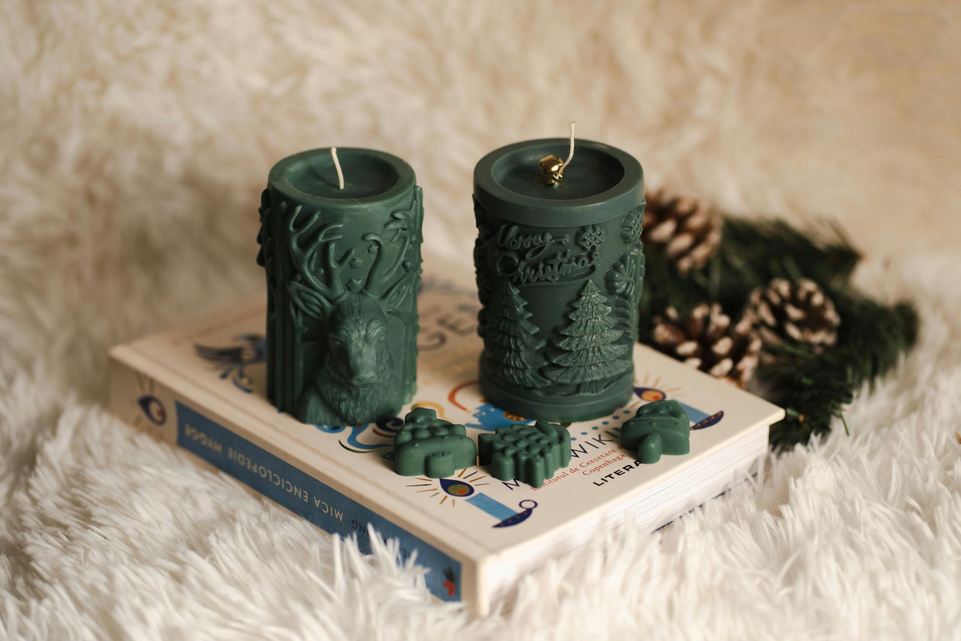 Two decorative green Christmas candles, one featuring a reindeer design and the other embossed with pine trees and "Merry Christmas," sit on a book with small festive wax shapes in front. Pinecones and evergreen branches in the background add a seasonal touch, making the image ideal for blogs about home staging, creating a cosy festive atmosphere, or selling property during the Christmas season.