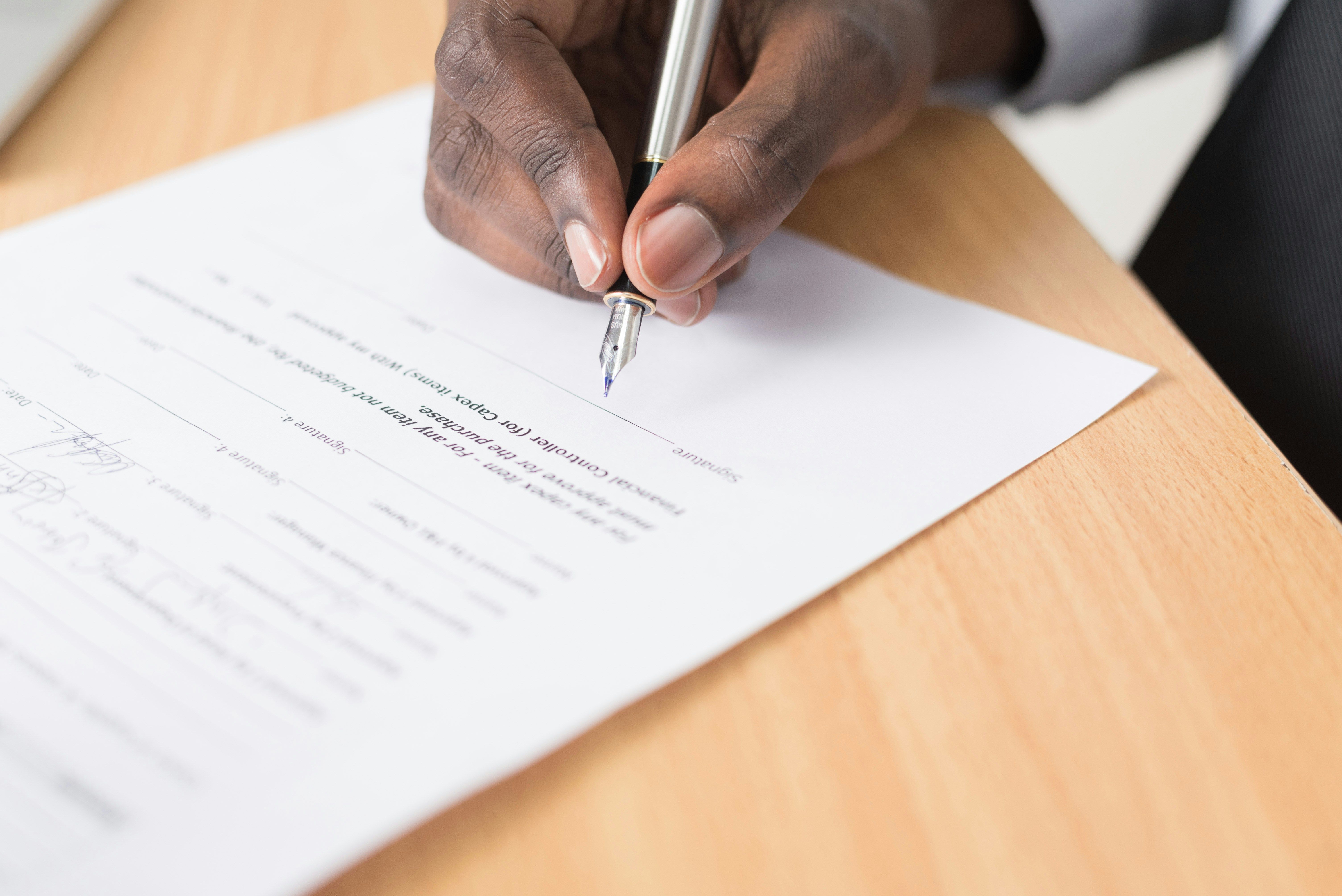 A professional close-up photograph of a person’s hand holding a fountain pen while signing a legal document on a wooden desk. Ideal for representing property contracts, agreements, probate paperwork, or signing on the dotted line with Integra Estates.