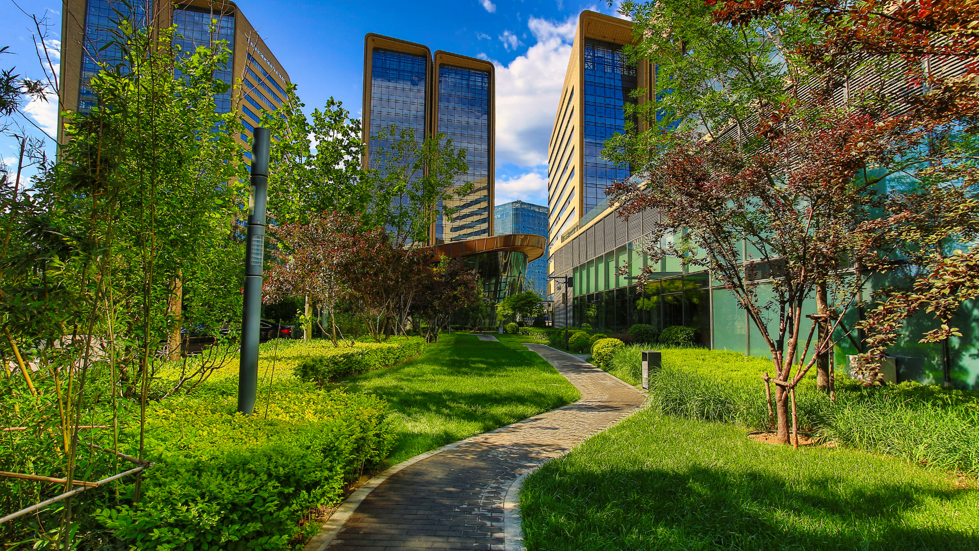 A modern business or commercial district featuring tall glass buildings surrounded by lush green landscaping. A winding stone pathway leads through well-maintained grass, bushes, and trees, creating a blend of nature and contemporary architecture under a bright blue sky with scattered clouds.