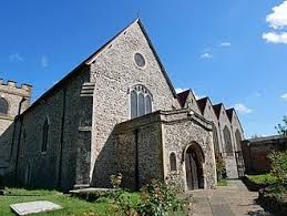 A historic stone church with a steep pitched roof and arched wooden entrance door, featuring tall narrow windows with decorative tracery. The exterior is built from irregular light-colored stones, giving it a rustic, medieval appearance. A clear blue sky forms the backdrop, and a grassy churchyard with a paved path surrounds the building.