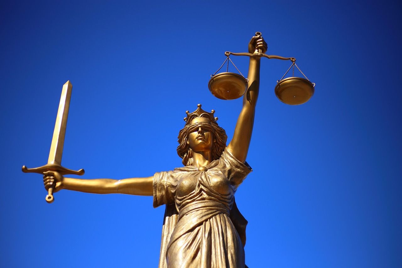 A golden statue of Lady Justice stands against a clear blue sky, holding a sword in her right hand and balanced scales in her left, symbolising fairness, law, and equality within the justice system.
