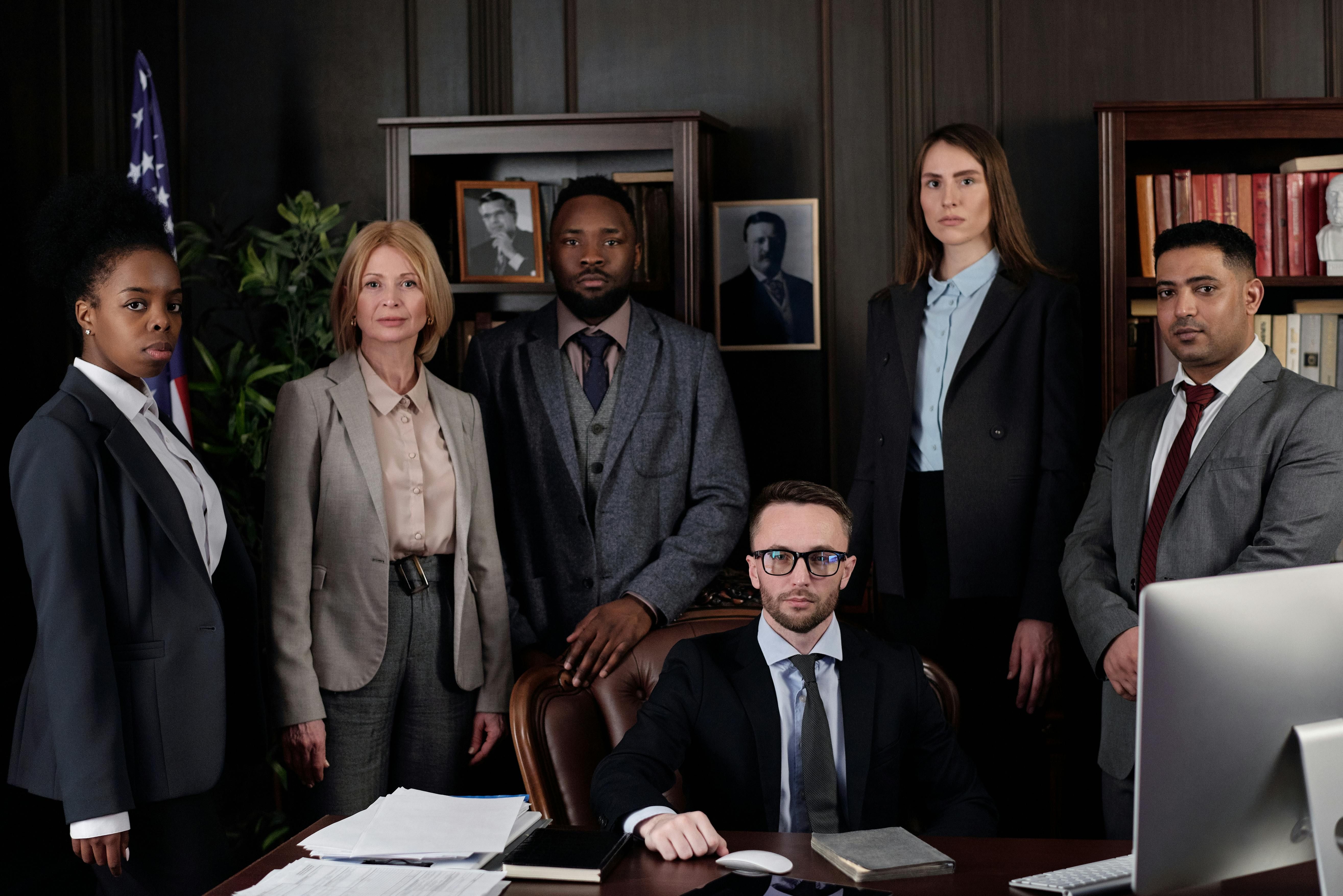 A diverse group of five professionally dressed people stand confidently in an office with dark wood furnishings and bookshelves, while one man sits at a desk with papers, books, and a computer in front of him. The formal setting and their serious expressions suggest a legal or business team portrait.