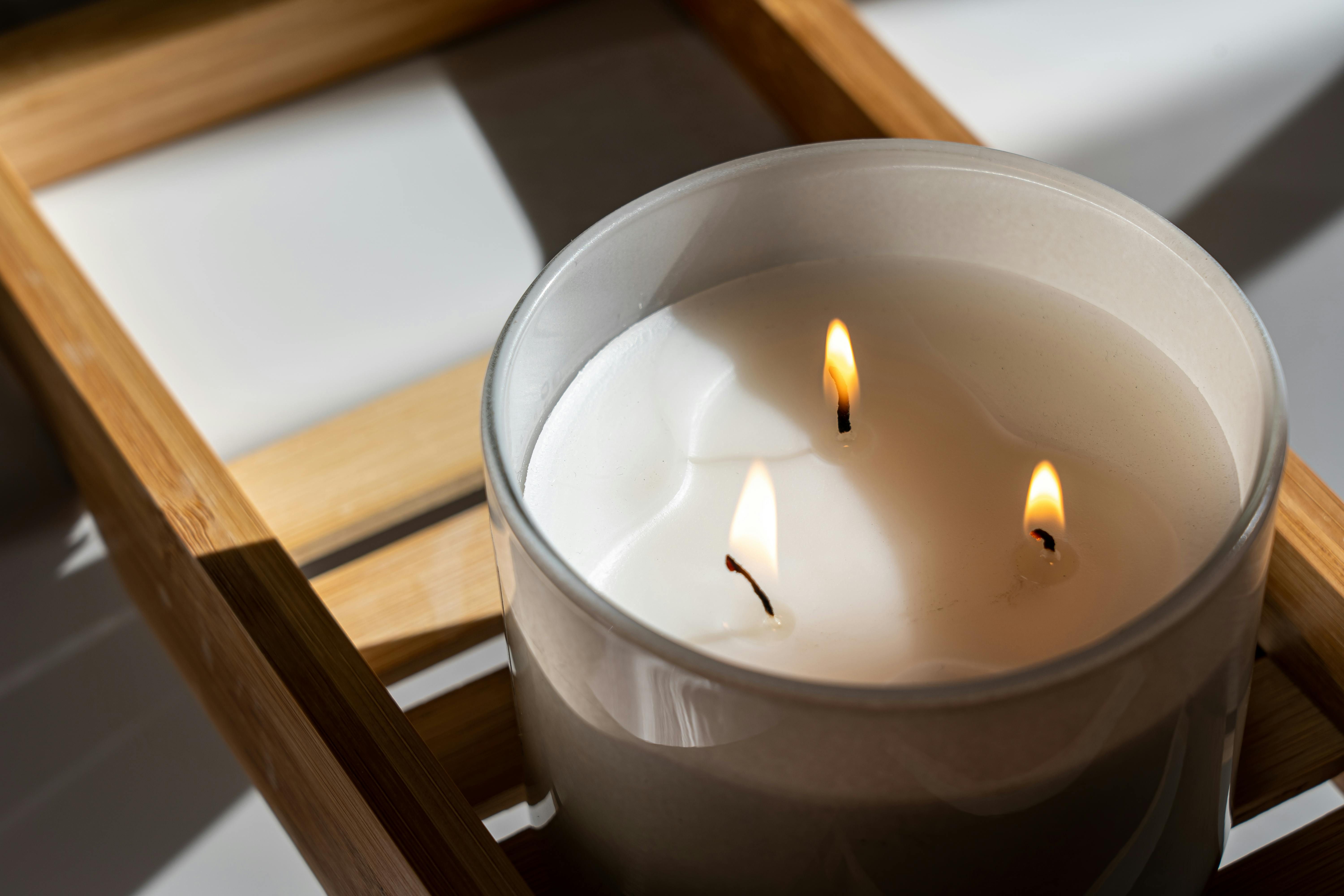A close-up of a lit three-wick white candle in a glass container placed on a wooden tray. The soft glow from the small flames casts gentle shadows on the surrounding surface, creating a warm and calming atmosphere.