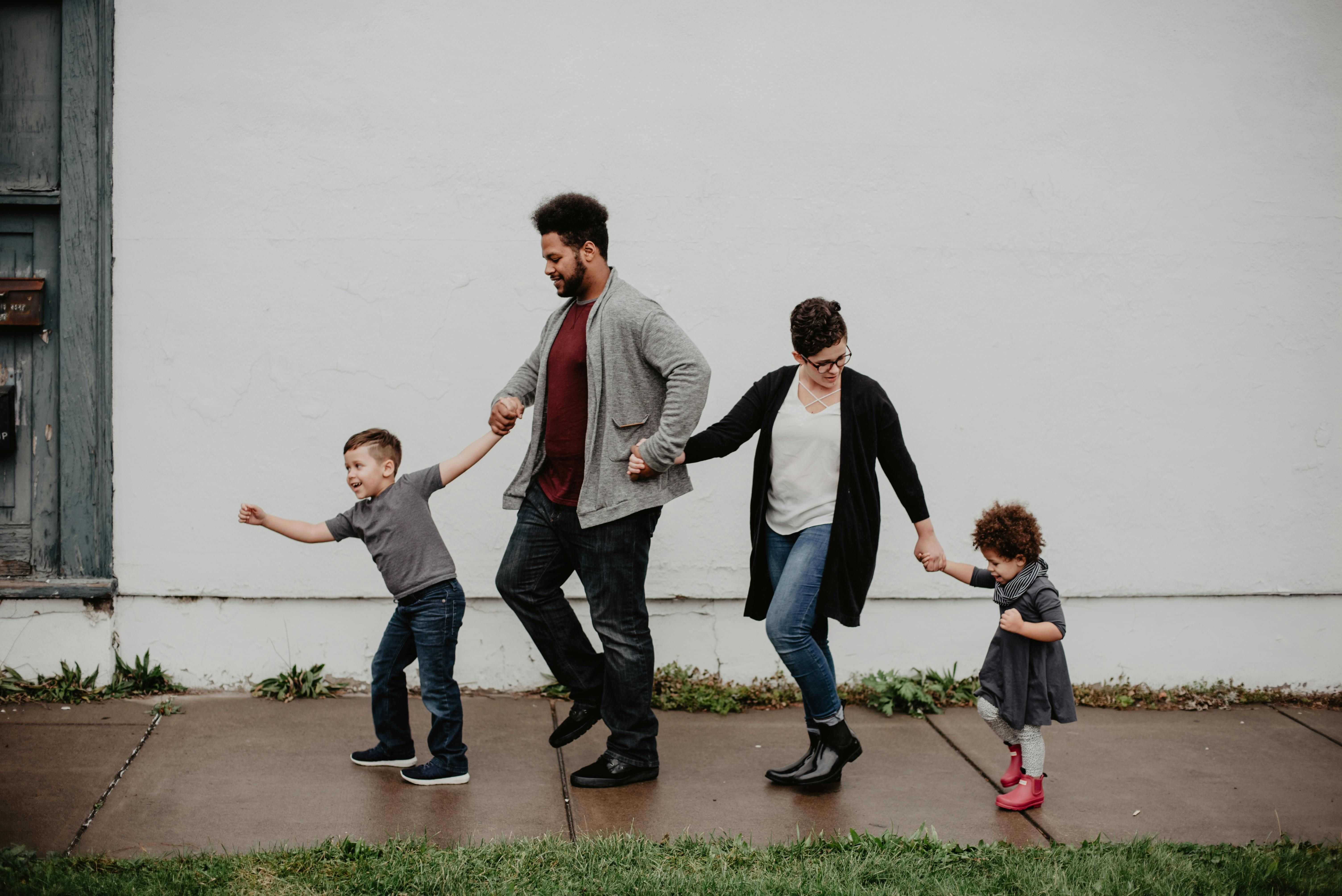 A cheerful family of four walking hand in hand along a sidewalk in front of a white wall. The father and mother are in the middle, holding hands with their two children a playful young boy pulling forward with excitement on one side, and a little girl in a gray dress and red boots on the other.