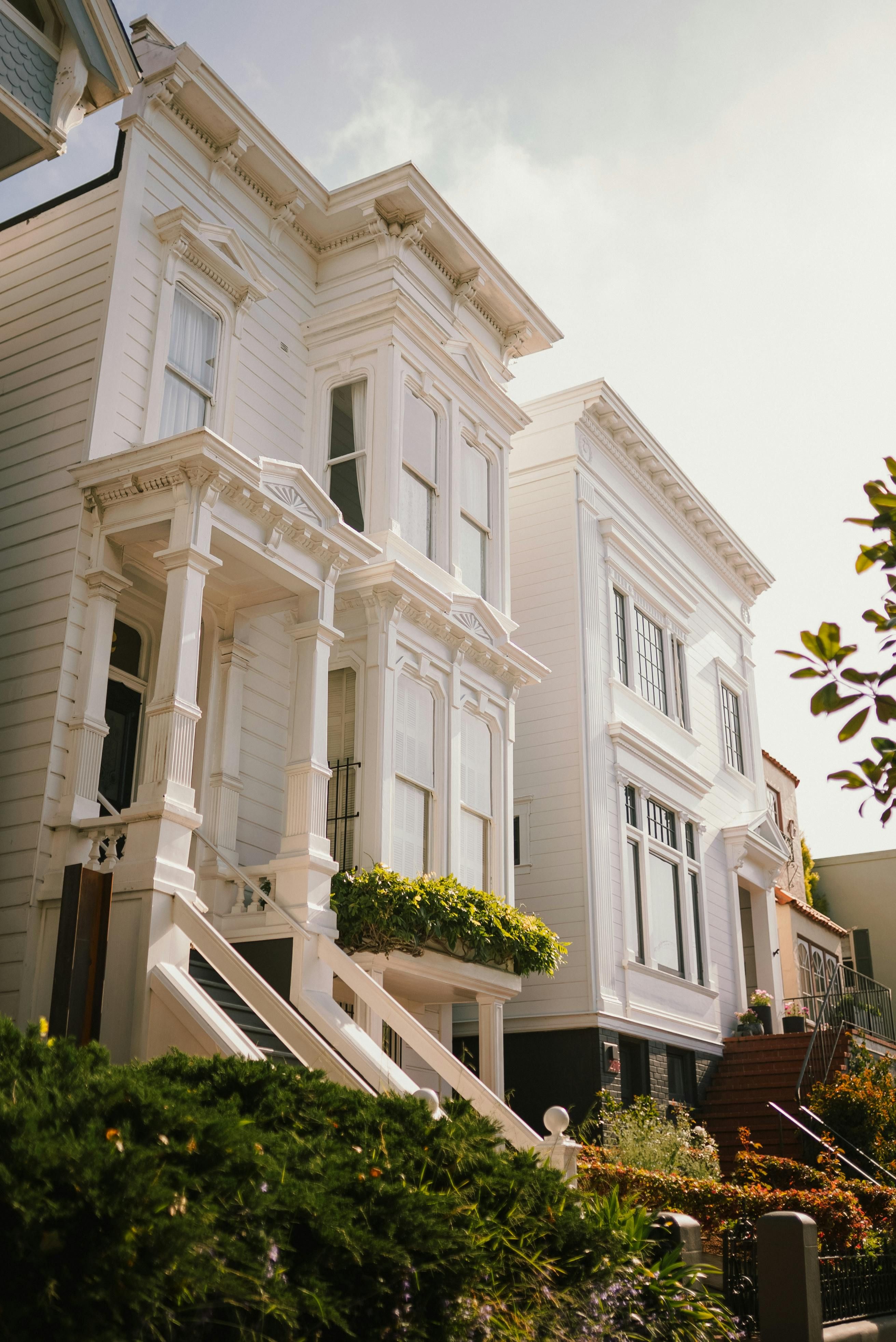 A bright, elegant row of Victorian-style houses painted in crisp white, featuring tall, narrow windows, ornate cornices, and decorative columns around the front porches. The homes stand on a slightly elevated entrance with stairs leading up, and a lush green hedge and garden sit in the foreground. Soft sunlight illuminates the facades, giving the scene a warm, inviting atmosphere.