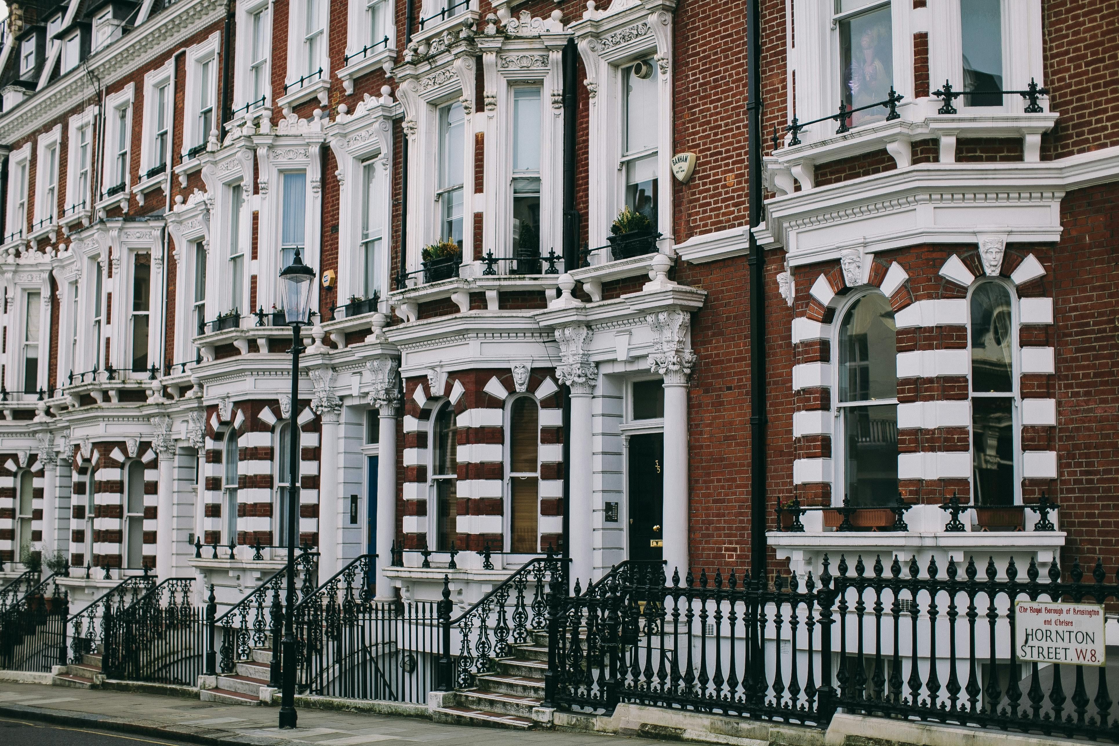 A row of elegant Victorian-style terraced houses on Hornton Street in Kensington, London. The buildings feature red brick facades with ornate white stone detailing, tall arched windows, decorative columns, and wrought-iron railings along the steps and balconies.