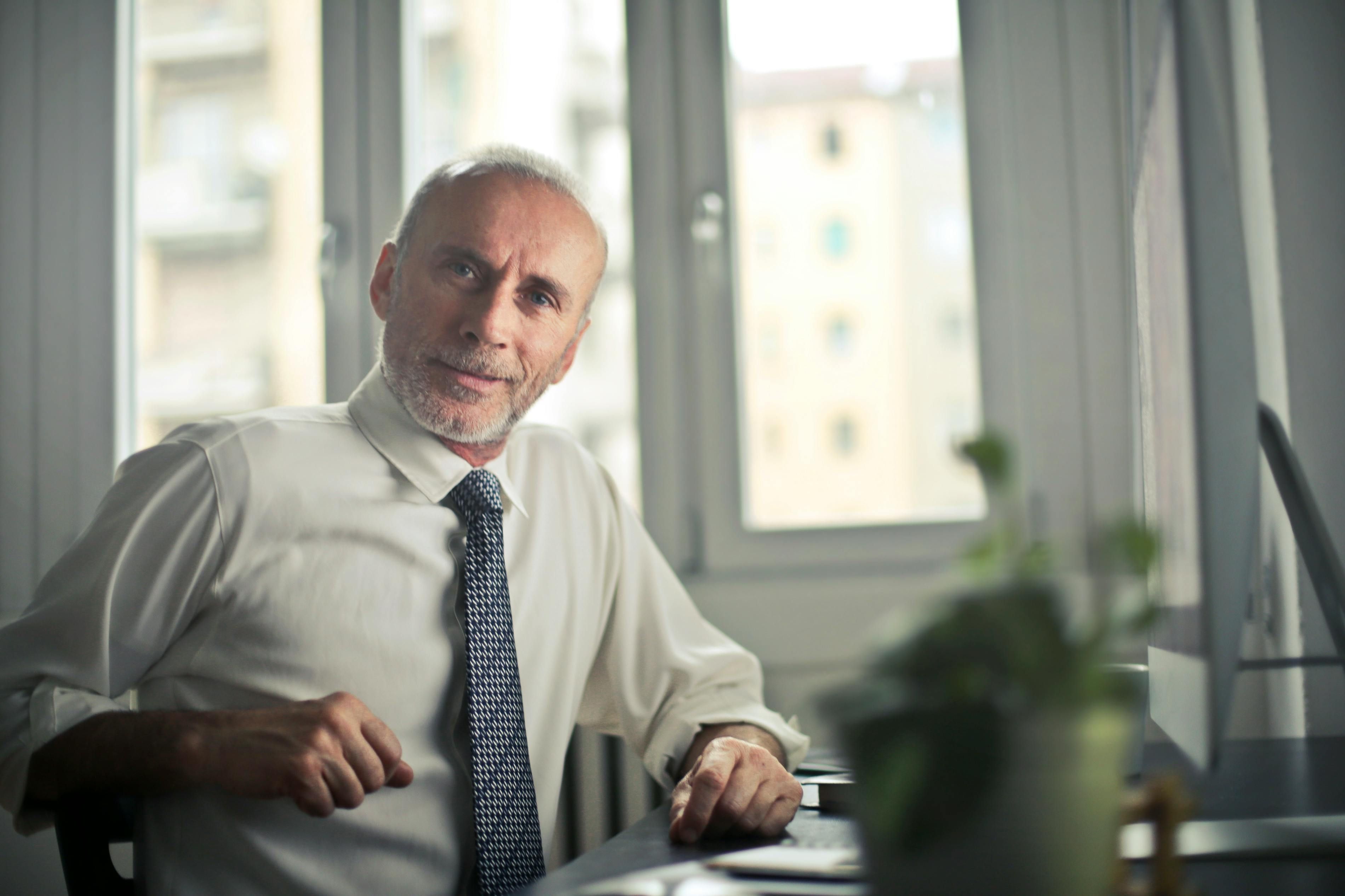 Confident mature businessman sitting at his desk in an office, wearing a white shirt and tie, with natural light coming through large windows in the background.