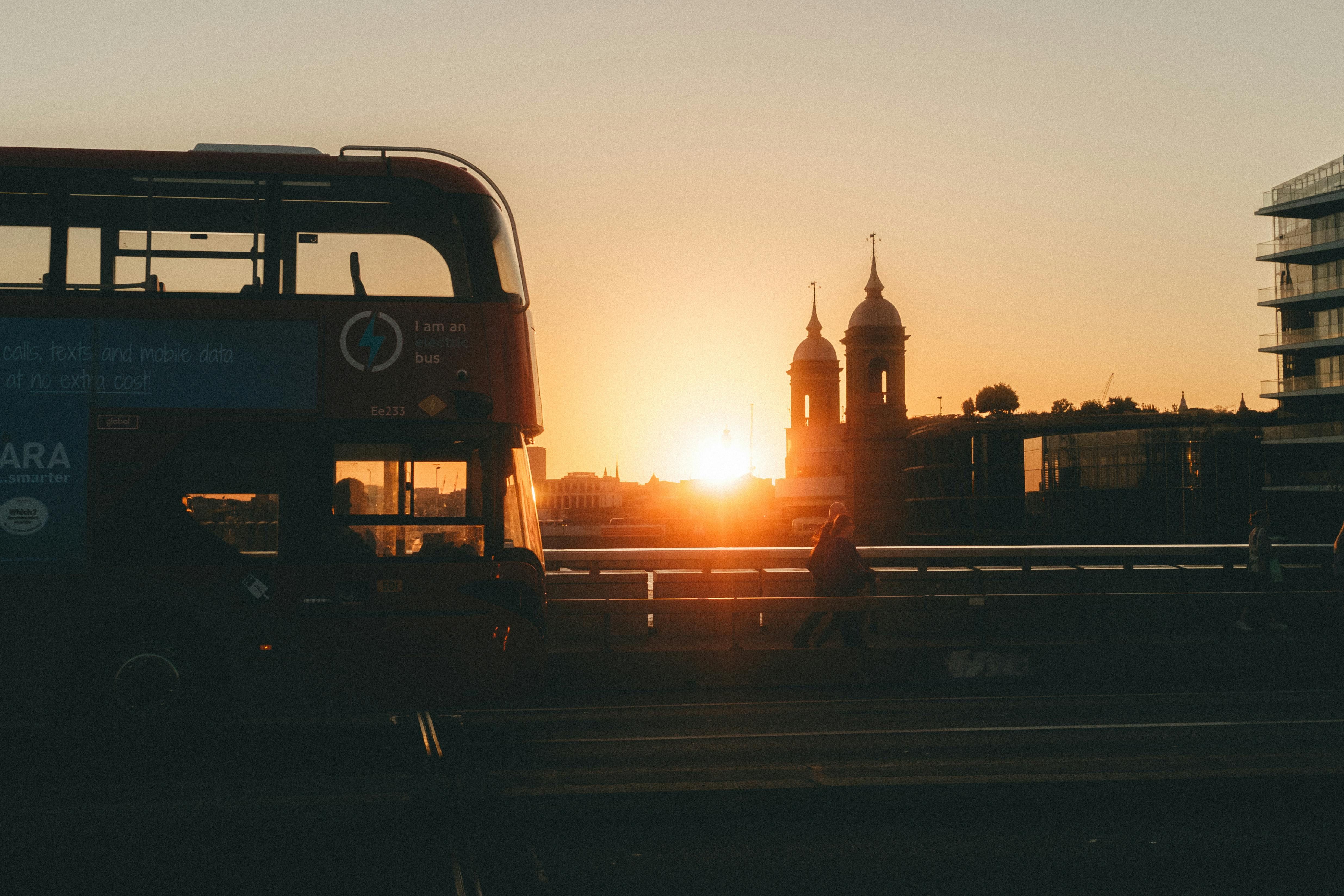 A red double-decker bus crosses a bridge at sunset, silhouetted against the golden sky. The sun sets behind twin-domed towers in the distance, casting a warm glow over the cityscape. Pedestrians walk along the bridge while modern and historic buildings frame the horizon, capturing the calm and beauty of an evening in London.